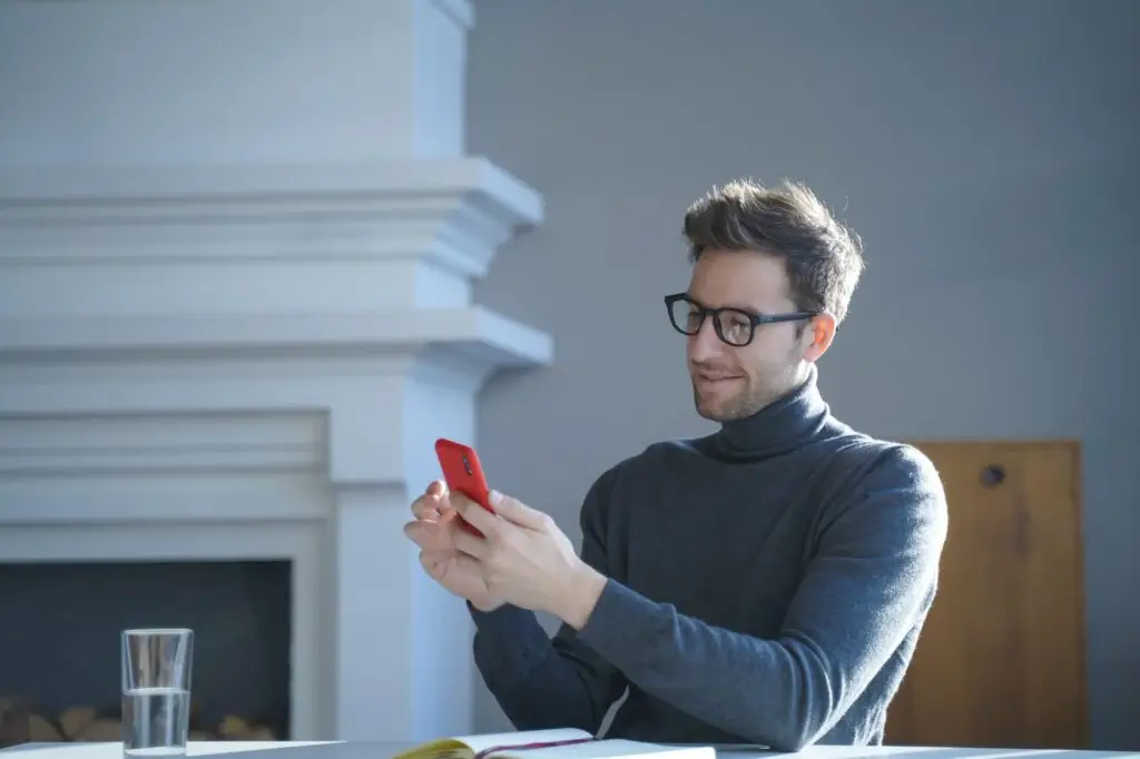 Young handsome german man in glasses sitting at table and using mobile phone at home
